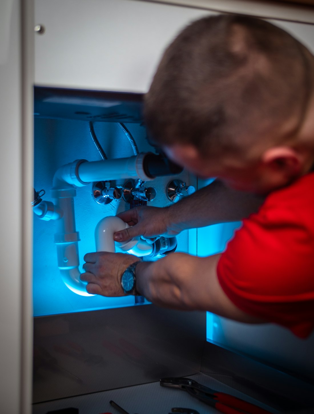 A plumber kneeling at an open cabinet, working on the pipes under a sink